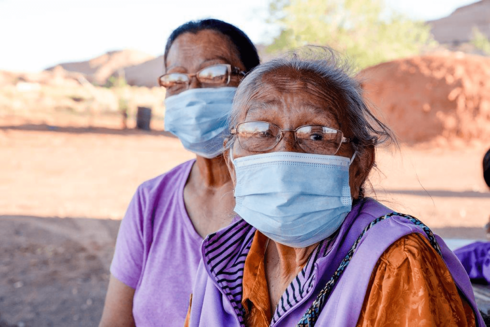 An elderly indigenous woman and a middle-aged indigenous woman both wearing glasses and masks sitting together outside in a dessert environment.