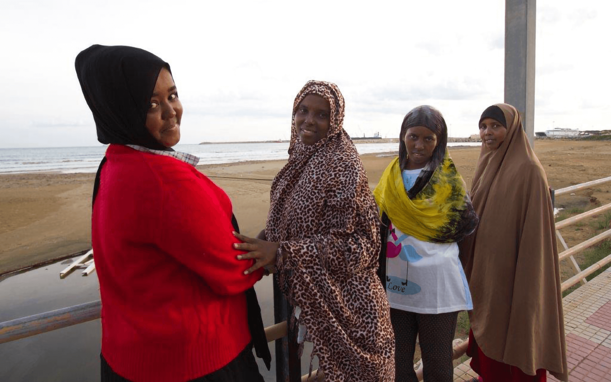 Four female Muslim women wearing hijabs standing on a boardwalk by the ocean, looking back and smiling.